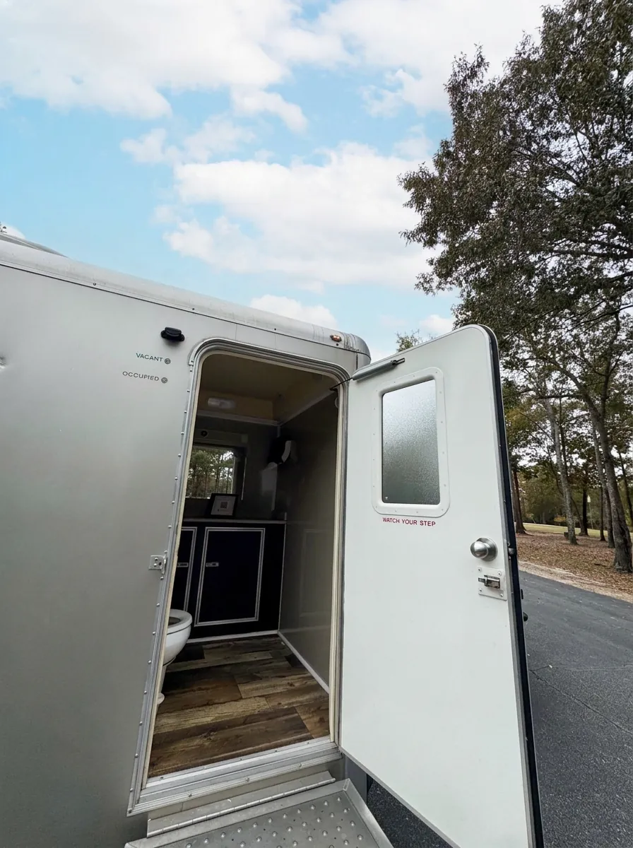 Open door of an Augusta VIPee restroom trailer showing the wood-look floor, vanity and flushing toilet